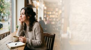 Young woman looking thoughtfully out a cafe window while doing a mid-year life review in her journal next to a cup of coffee.